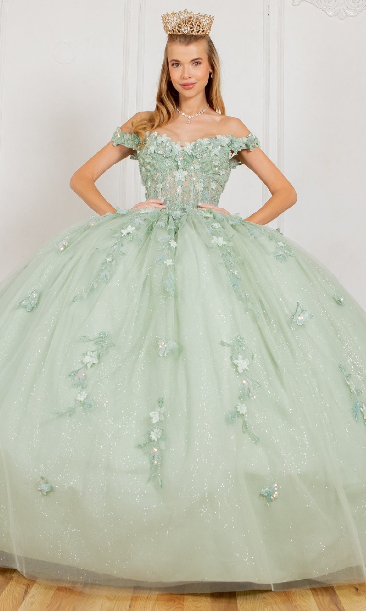 A woman stands crowned with hands on her waist, wearing the Cinderella Couture 8075J quinceanera dress—light green, off-the-shoulder, adorned with floral details and 3D appliques.